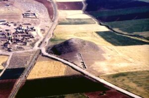 Aerial view of patchwork fields with a winding road and a small village on the left, plus a large rounded hill with a row of rectangular structures along its slope.