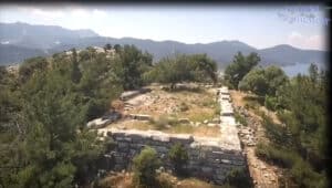 Stone terrace ruins on a hillside with shrubs and trees, mountains and a lake visible in the distance.