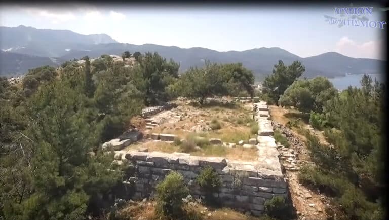 Stone terrace ruins on a hillside with shrubs and trees, mountains and a lake visible in the distance.