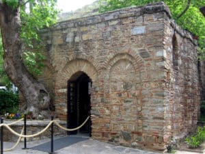 Stone brick historic building with a rounded arched doorway, open black door, and a rope barrier in front amid trees and flowers.
