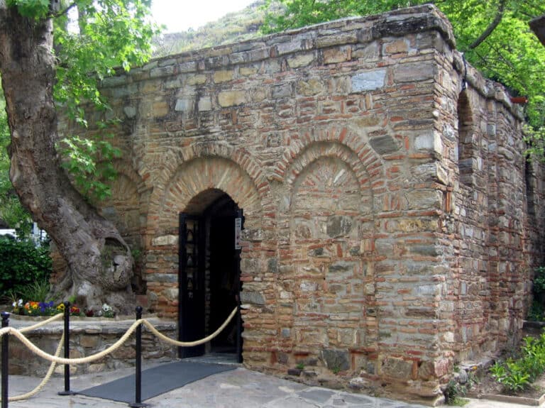 Stone brick historic building with a rounded arched doorway, open black door, and a rope barrier in front amid trees and flowers.