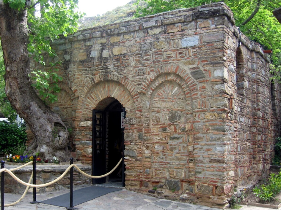 Stone brick historic building with a rounded arched doorway, open black door, and a rope barrier in front amid trees and flowers.