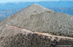 Dirt road winds along a rocky hillside with distant blue mountains and wind turbines on the ridge
