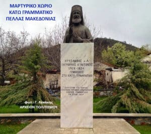 Monument with a tall marble pedestal and statue on top in a rural Greek village square, with Greek inscriptions on the pedestal and surrounding trees in the background.