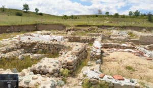 Archaeological excavation site with low stone walls, exposed earth and scattered red tiles, set in a grassy field under a blue sky.