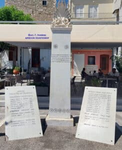 White marble monument with a sculpted flame top, flanked by two Greek-inscribed plaques; outdoor cafe seating visible behind.