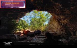 Inside a dark cave looking toward a sunlit exit with trees; a sign in the upper‑left reads 'Skotino Cave' (Greek/English).