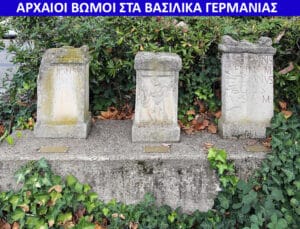 Three weathered stone monuments with carvings sit on a raised concrete platform, surrounded by ivy and fallen leaves in a garden setting.