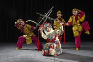Performers in colorful Chinese opera costumes with ornate masks on stage, crossing shiny swords in a dynamic pose.
