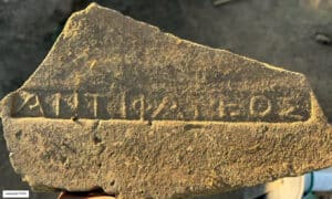 Fragment of a weathered stone with a carved line of capital letters along a shallow groove; the text is faint and partially eroded.