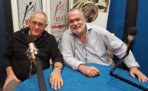 Two men sit at a blue table in a radio studio with large microphones and a Kriti FM banner behind them, smiling at the camera.