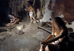 Scene of a cave-dwelling group: a shirtless man with a spear stands by a seated figure near a fire, while others work on hides and tools around them in a rough, dirt floor shelter.
