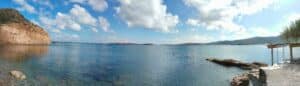 Panoramic view of a calm turquoise bay with a rocky cliff on the left, distant islands, and a rocky shoreline on the right.