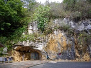 Rocky hillside with a cave entrance; vegetation on top and a small seating area near the opening with a person nearby.