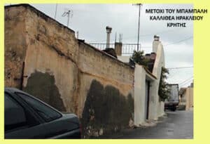 Narrow street with a tall weathered wall on the left and a car parked in the foreground; Greek text appears in the upper right, suggesting a location in Crete.