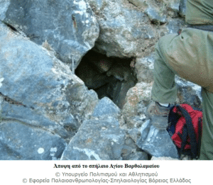 Hiker in green pants and gloves steps toward a dark cave entrance among rocky terrain, with a red backpack nearby.