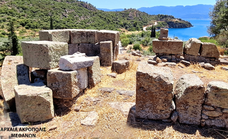 Ancient stone ruin with large rectangular blocks on a dry hillside, overlooking a blue sea; Greek text in the lower-left corner.