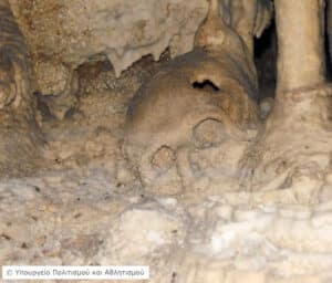 Cave interior with mineral formations and a rounded, hollow rock on the floor amid stalagmites and stalactites.