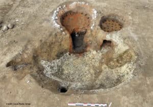 Two circular, rusty metal drainage covers set in dirt during an excavation, with mineral crust around them and a measurement scale nearby.