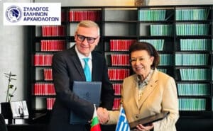 Two professionals shake hands in an office, with Greek and Bulgarian flags on the desk and shelves of red and green binders behind them, plus a logo in the corner.